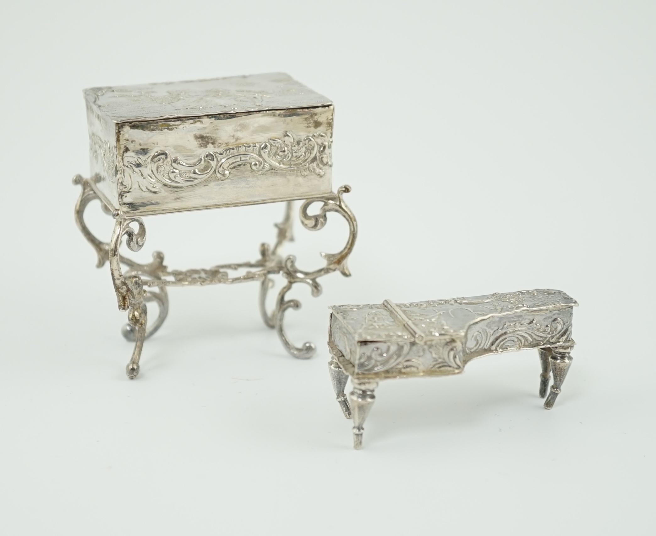 A late 19th century continental embossed silver miniature model of grand piano, length 53mm, together with a similar model of a table/desk on ornate scroll supports